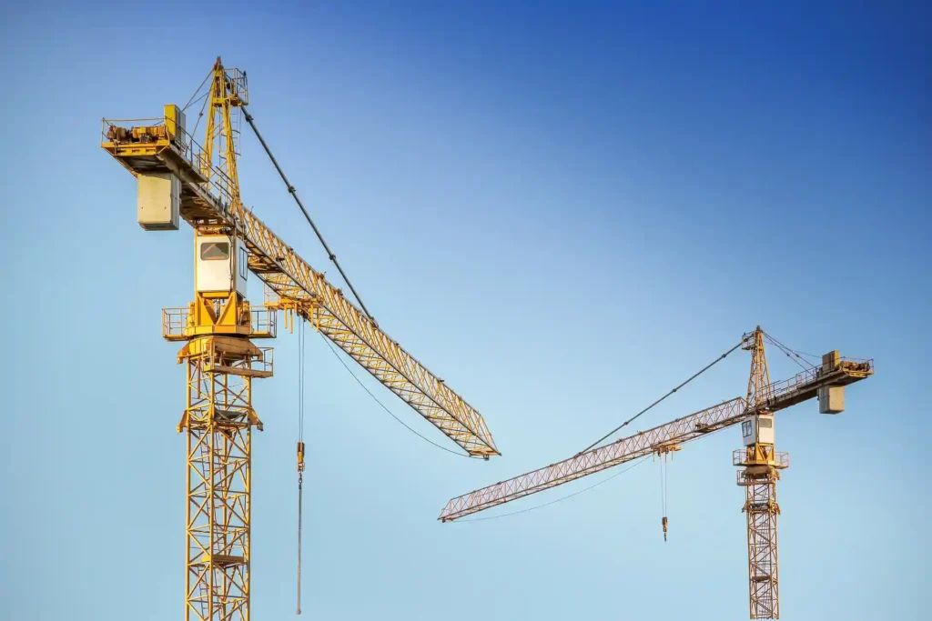 Two large construction cranes against a clear blue sky, representing tools used in construction project management for lifting and building large structures.