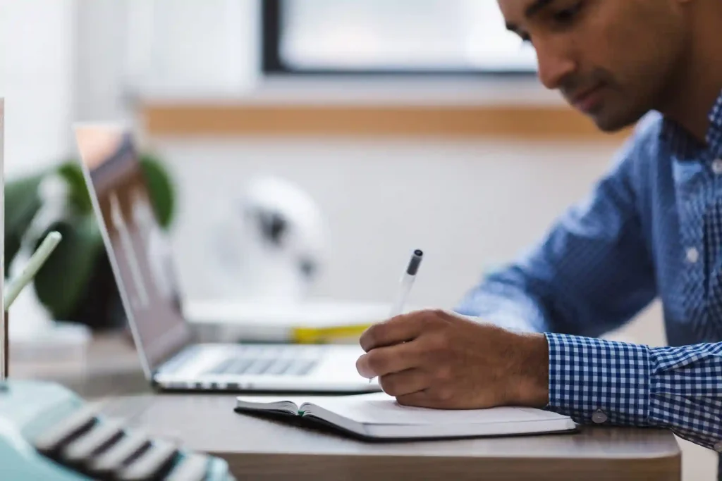 Architect conducting a feasibility study, making notes with a pen while working on a laptop, representing the initial analysis and planning phase in architecture.